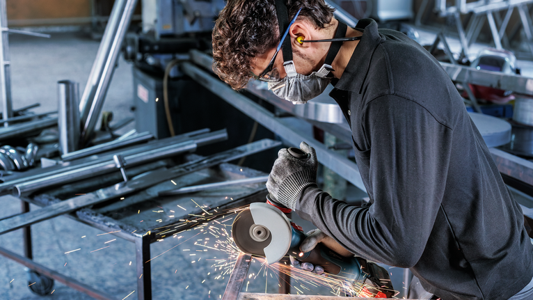 Orang yang mengenakan peralatan keselamatan menggunakan penggiling sudut untuk memotong logam, menghasilkan percikan api.