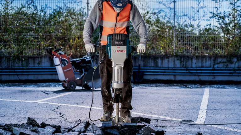 Seseorang yang mengenakan perlengkapan keselamatan menggunakan palu pembongkaran untuk menghancurkan perkerasan beton.