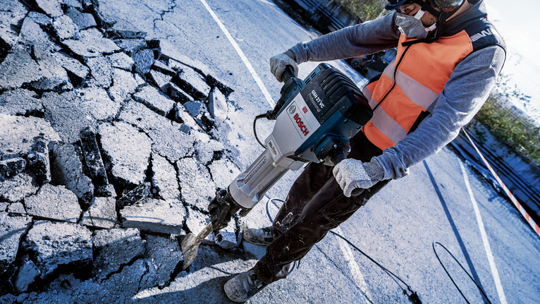 Seseorang yang mengenakan peralatan keselamatan memecah aspal dengan palu pembongkaran.