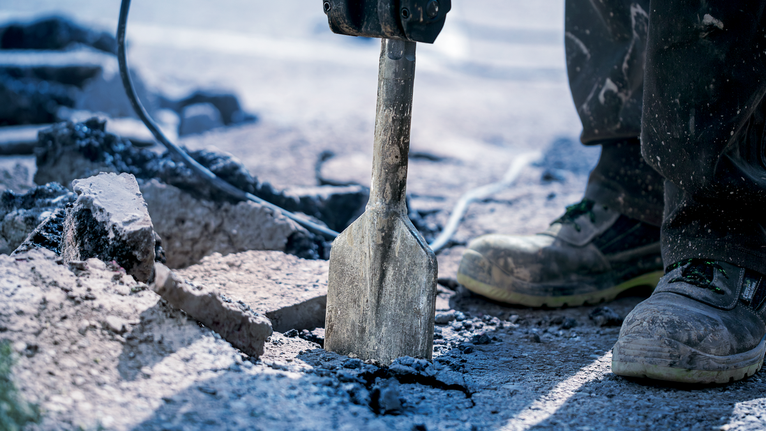 Seseorang yang mengenakan peralatan keselamatan memecahkan beton dengan alat pembongkaran.