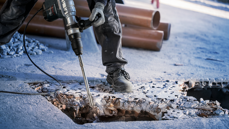 Orang yang mengenakan peralatan keselamatan sedang memecahkan lantai beton dengan palu pembongkaran.