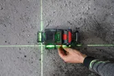 Seseorang sedang mengatur alat perata laser yang memproyeksikan garis hijau pada lantai beton.