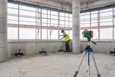 Orang yang mengenakan perlengkapan keselamatan sedang menyelaraskan material di dekat alat perata laser pada tripod.