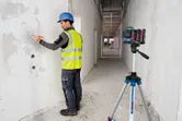 Seseorang yang mengenakan perlengkapan keselamatan menandai dinding dengan panduan alat perataan laser pada tripod.