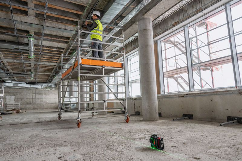 Seseorang yang mengenakan perlengkapan keselamatan bekerja di perancah, dengan alat perata laser yang memproyeksikan garis.