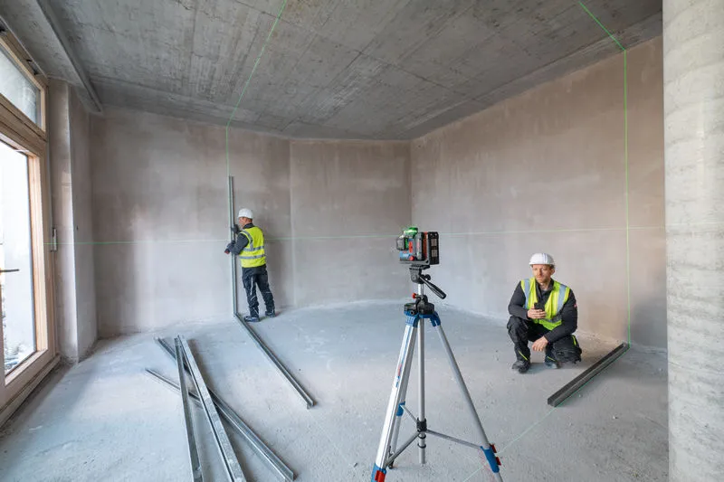 Dua orang yang mengenakan perlengkapan keselamatan menggunakan alat perata laser di ruangan yang belum selesai.