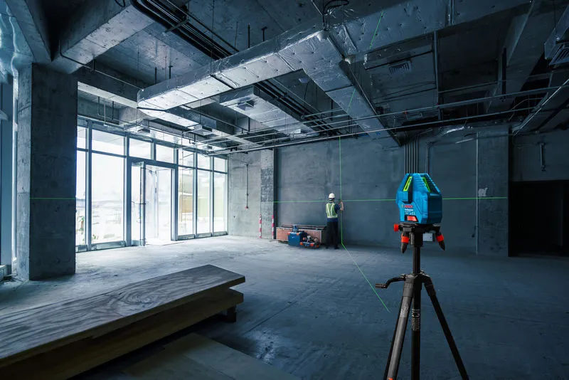 Seseorang yang mengenakan perlengkapan keselamatan menyelaraskan dinding menggunakan alat perata laser di lokasi konstruksi.