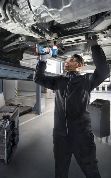 Seseorang yang mengenakan peralatan keselamatan menggunakan bor tanpa kabel di bawah kendaraan yang terangkat.