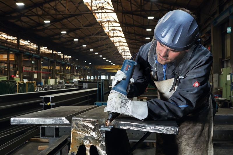 Seseorang yang mengenakan perlengkapan keselamatan menggunakan penggiling lurus pada balok logam.