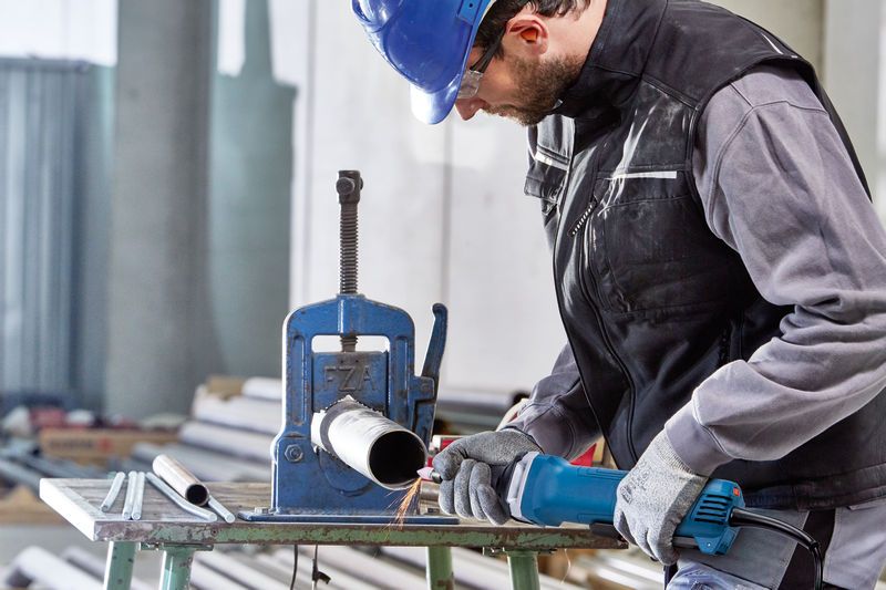 Seseorang yang mengenakan perlengkapan keselamatan menggunakan penggiling lurus untuk mengerjakan pipa logam.