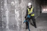Seseorang yang mengenakan peralatan keselamatan memecahkan beton dengan palu pembongkaran tanpa kabel.