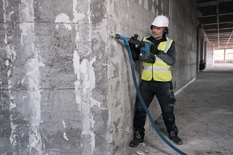 Seseorang yang mengenakan perlengkapan keselamatan menggunakan palu pembongkaran tanpa kabel pada dinding beton.