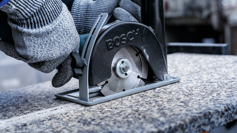 Seseorang yang mengenakan perlengkapan keselamatan menggunakan gergaji logam untuk memotong lempengan granit.