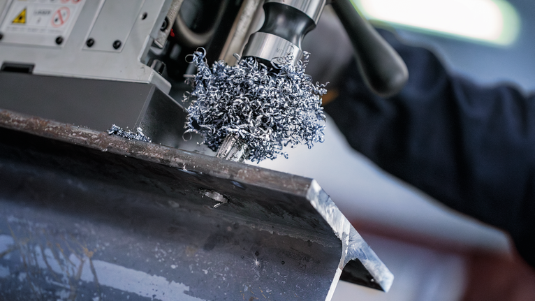 Orang yang mengenakan peralatan keselamatan mengebor balok logam, sehingga menghasilkan serutan logam.