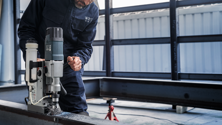 Seseorang yang mengenakan peralatan keselamatan mengoperasikan bor magnet pada balok baja.