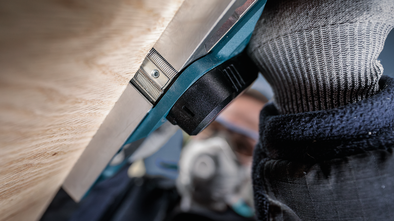 Orang yang mengenakan peralatan keselamatan mengoperasikan perkakas listrik di dekat permukaan kayu.