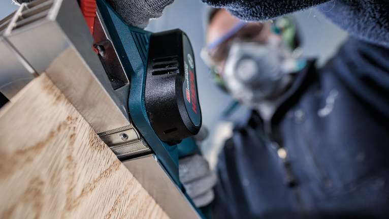 Orang yang mengenakan perlengkapan keselamatan menggunakan alat serut listrik pada papan kayu.