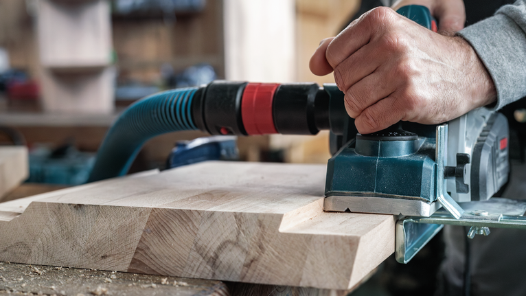Seseorang menggunakan alat serut listrik pada papan kayu di bengkel.