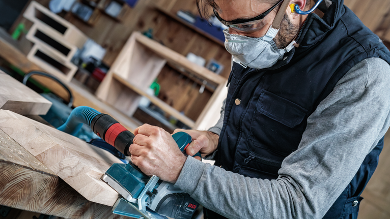 Orang yang mengenakan peralatan keselamatan mengoperasikan gergaji listrik untuk memotong papan kayu.