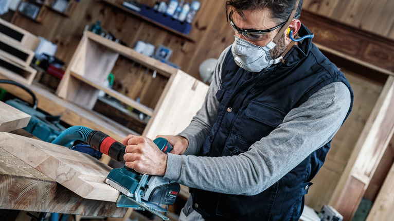 Orang yang mengenakan perlengkapan keselamatan menggunakan planer listrik untuk menghaluskan papan kayu.