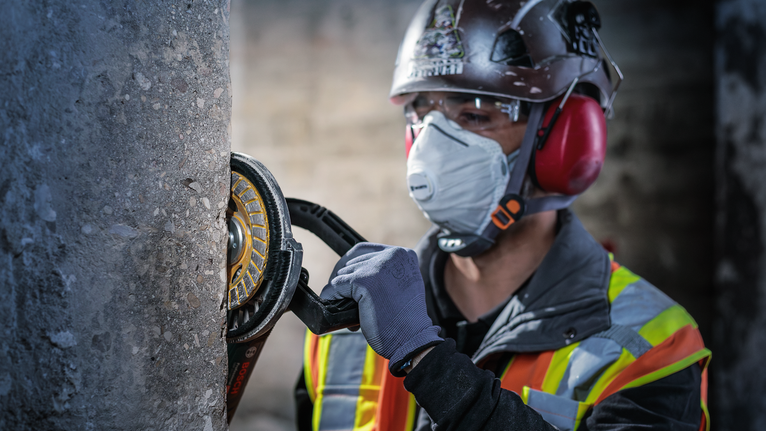 Seseorang yang mengenakan perlengkapan keselamatan menggiling pilar beton dengan perkakas listrik.
