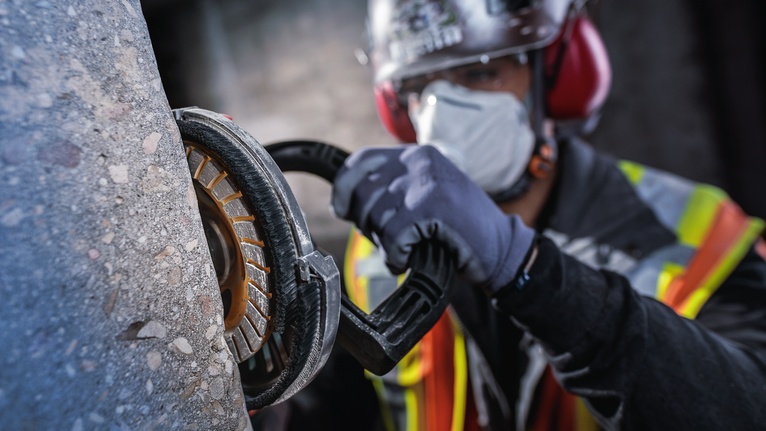 Orang yang mengenakan peralatan keselamatan menggiling beton dengan alat listrik.