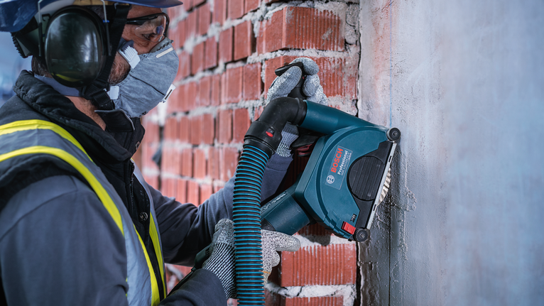 Pekerja yang mengenakan perlengkapan keselamatan menggunakan perkakas listrik untuk menggiling mortar dari dinding bata.