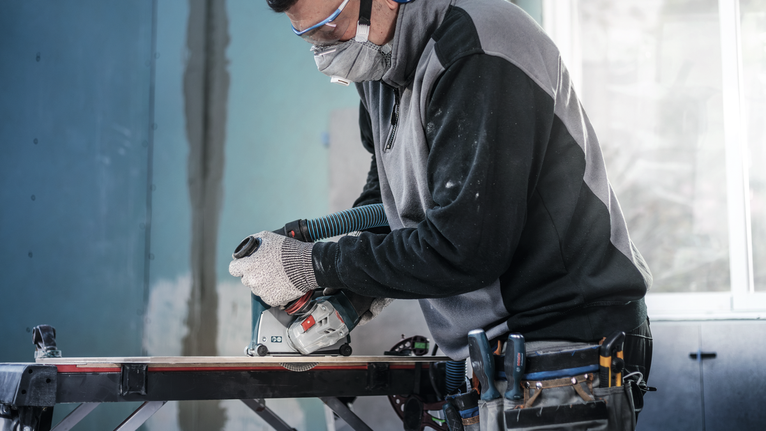 Orang yang mengenakan peralatan keselamatan menggunakan gergaji bundar untuk memotong kayu di meja kerja.
