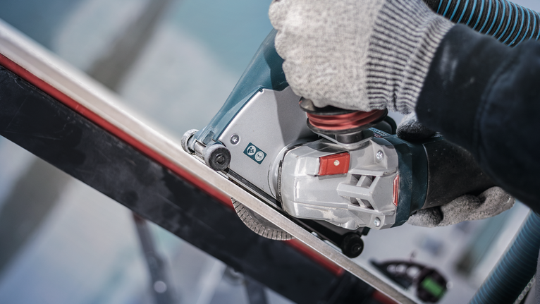 Orang yang mengenakan perlengkapan keselamatan menggunakan alat listrik untuk memotong batang logam.