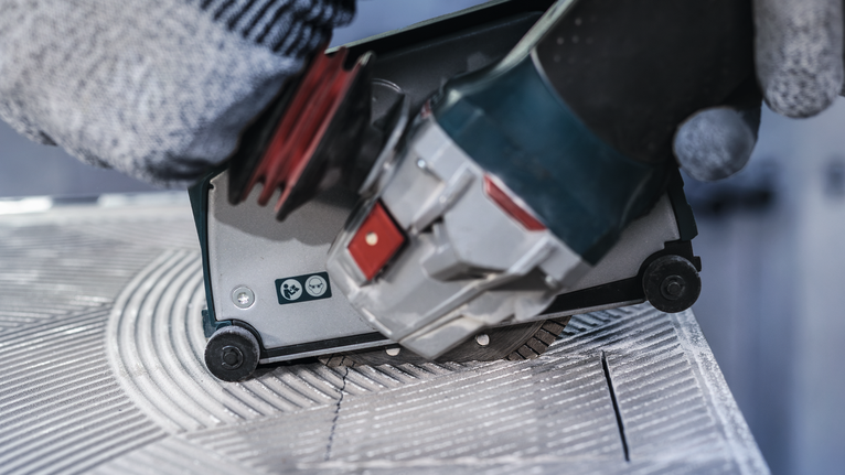 Orang yang mengenakan peralatan keselamatan memandu alat listrik untuk memotong alur pada ubin.