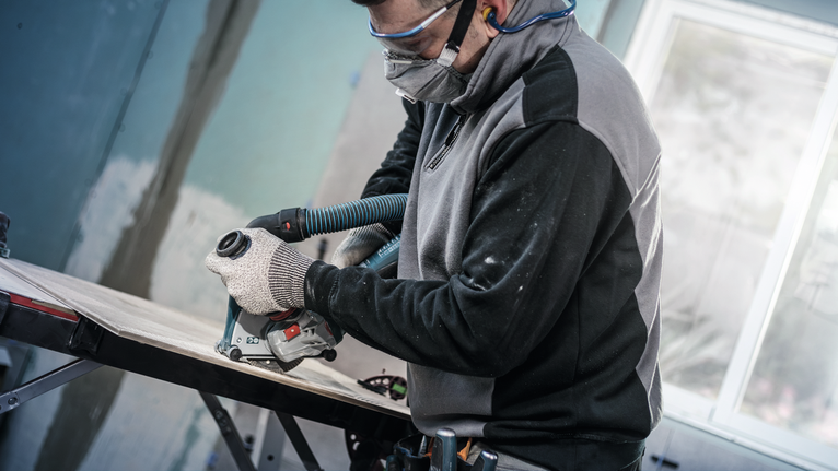 Orang yang mengenakan peralatan keselamatan memotong kayu dengan alat listrik yang dihubungkan ke selang vakum.