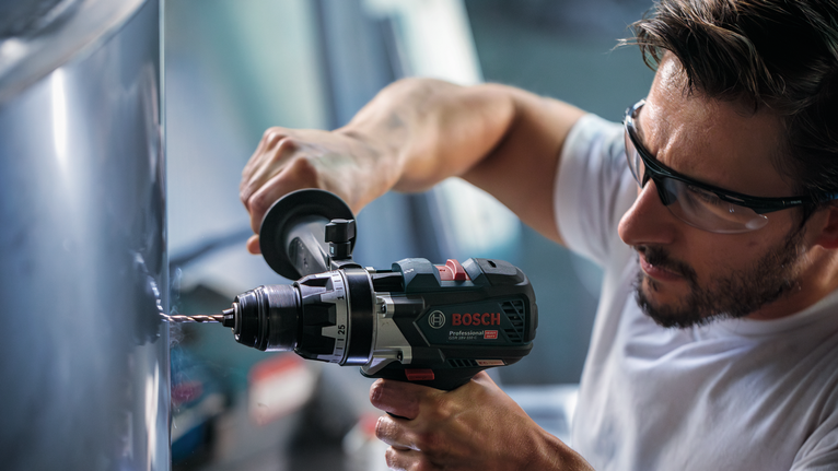 Seseorang yang mengenakan peralatan keselamatan mengebor logam dengan alat listrik.