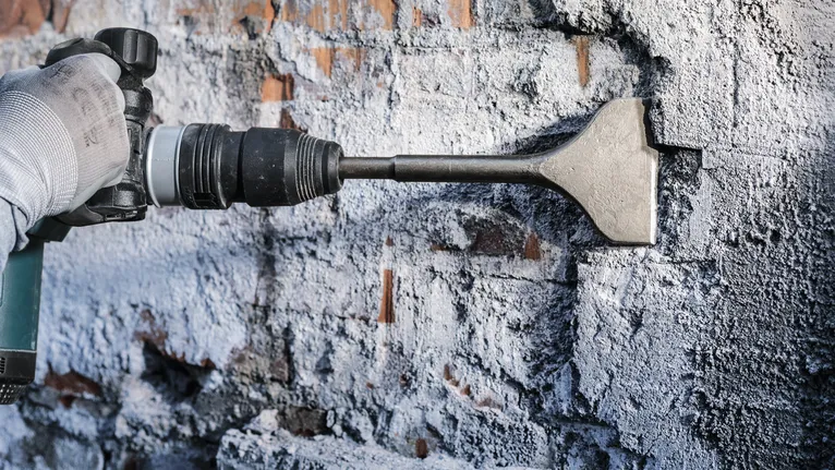 Orang yang mengenakan peralatan keselamatan menggunakan bor pahat untuk menghilangkan plester dari dinding bata.