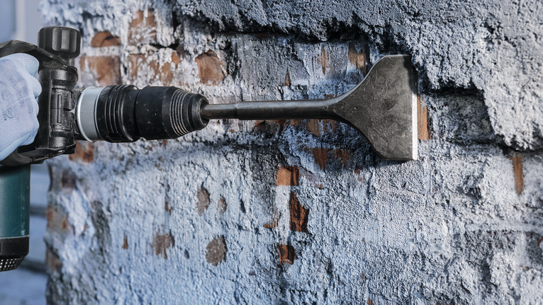 Orang yang mengenakan perlengkapan keselamatan menggunakan alat pahat untuk menghilangkan plester dari dinding.