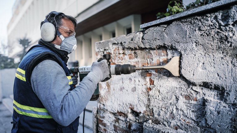 Orang yang mengenakan perlengkapan keselamatan menggunakan pahat listrik untuk menghilangkan mortar dari dinding bata.
