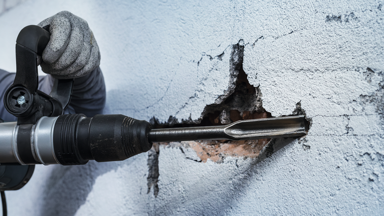 Seseorang yang mengenakan perlengkapan keselamatan menggunakan palu putar untuk memahat dinding.