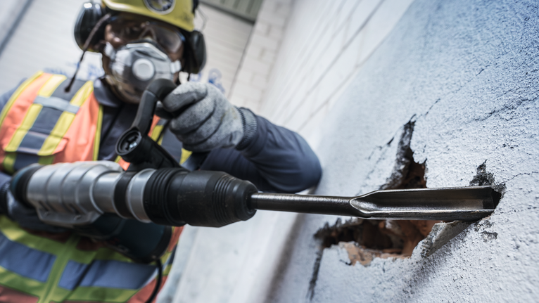 Orang yang mengenakan peralatan keselamatan menggunakan palu putar untuk menerobos dinding beton.