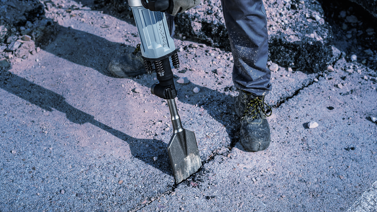 Seseorang yang mengenakan peralatan keselamatan memecahkan beton dengan palu pembongkaran.