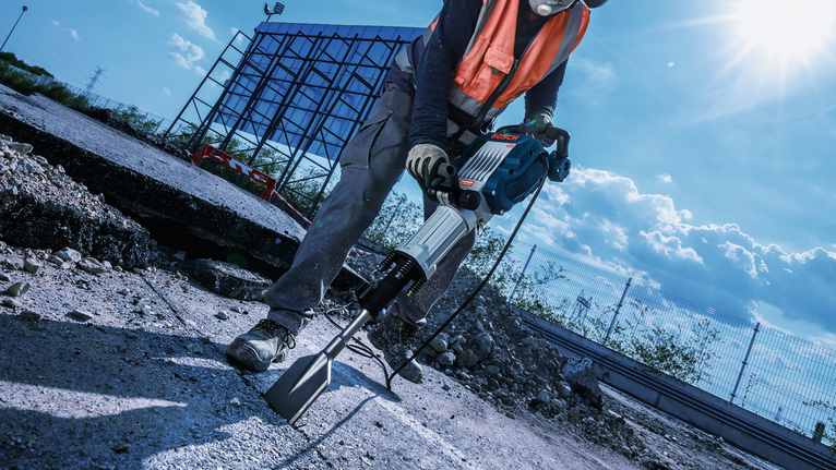 Seseorang yang mengenakan perlengkapan keselamatan menggunakan jackhammer untuk memecahkan beton di lokasi konstruksi.