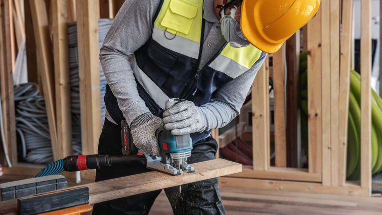 Seseorang yang mengenakan peralatan keselamatan memotong kayu dengan alat listrik di lokasi konstruksi.