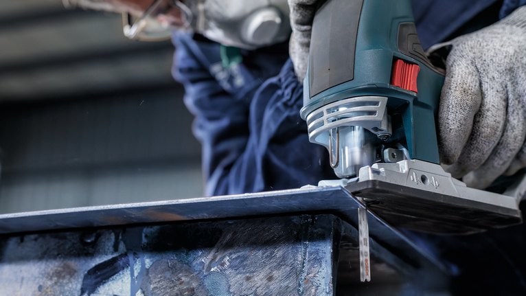 Orang yang mengenakan perlengkapan keselamatan menggunakan gergaji ukir untuk memotong lembaran logam.