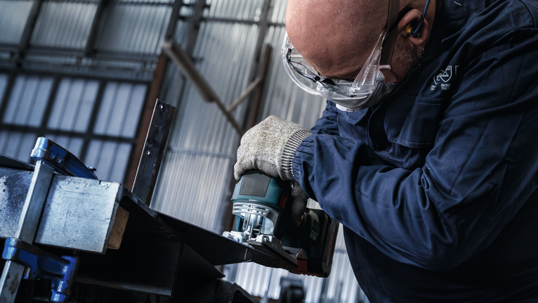 Orang yang mengenakan perlengkapan keselamatan menggerus tepi balok logam.