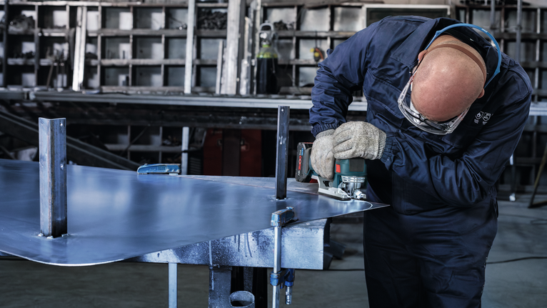Seseorang yang mengenakan peralatan keselamatan memotong lembaran logam dengan alat listrik.