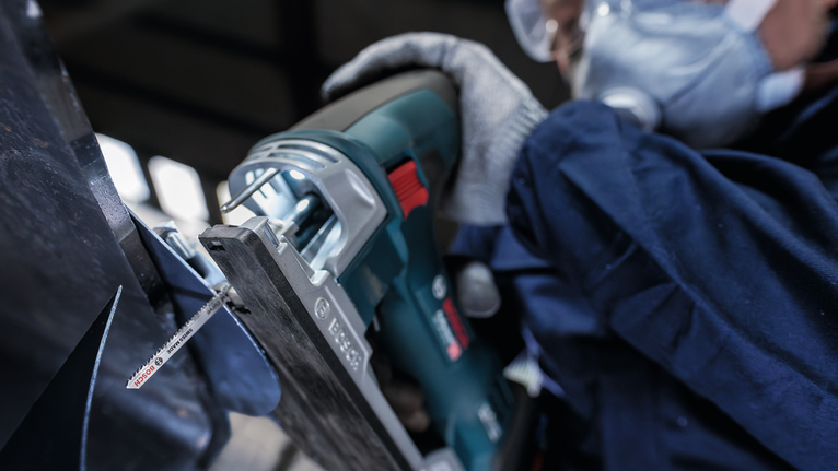 Orang yang mengenakan peralatan keselamatan memotong logam dengan gergaji bolak-balik.