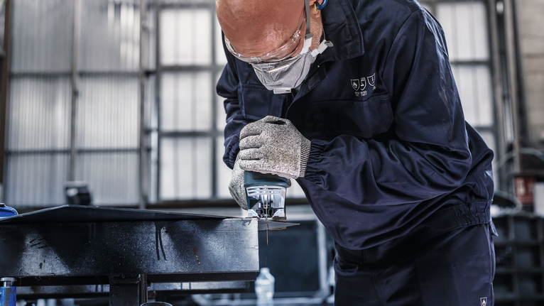 Orang yang mengenakan peralatan keselamatan memotong logam menggunakan gergaji listrik genggam.