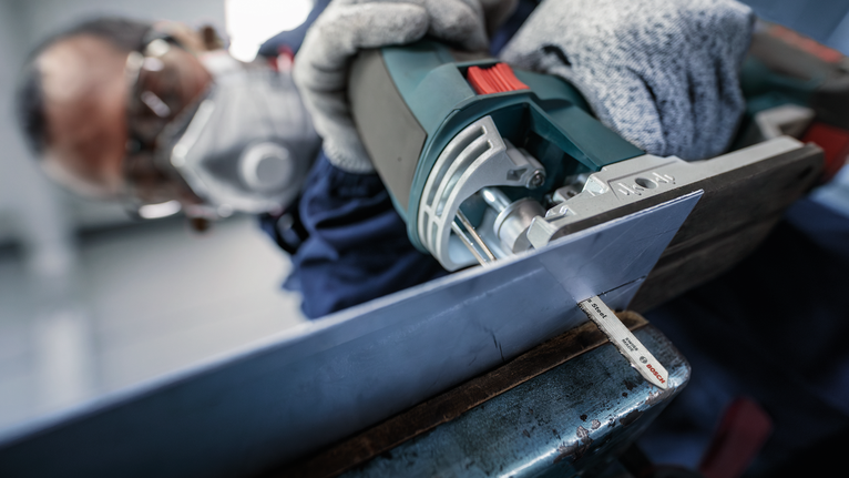 Seseorang yang mengenakan perlengkapan keselamatan menggunakan gergaji ukir untuk memotong lembaran logam.