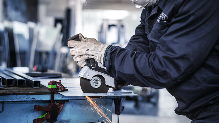 Orang yang mengenakan peralatan keselamatan menggunakan penggiling sudut untuk memotong logam, sehingga menimbulkan percikan api.