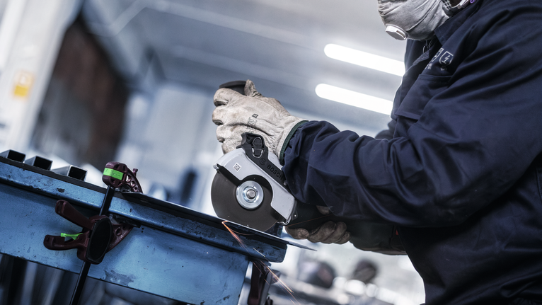 Orang yang mengenakan peralatan keselamatan menggiling logam dengan penggiling sudut, menghasilkan percikan api.