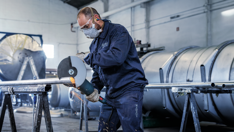 Seseorang yang mengenakan peralatan keselamatan menggiling pipa logam, sehingga menimbulkan percikan api.