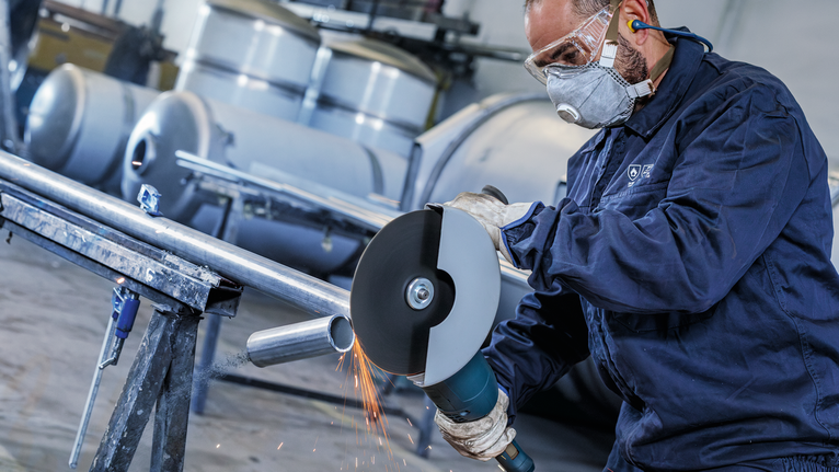 Seseorang yang mengenakan peralatan keselamatan memotong pipa logam dengan penggiling sudut besar.